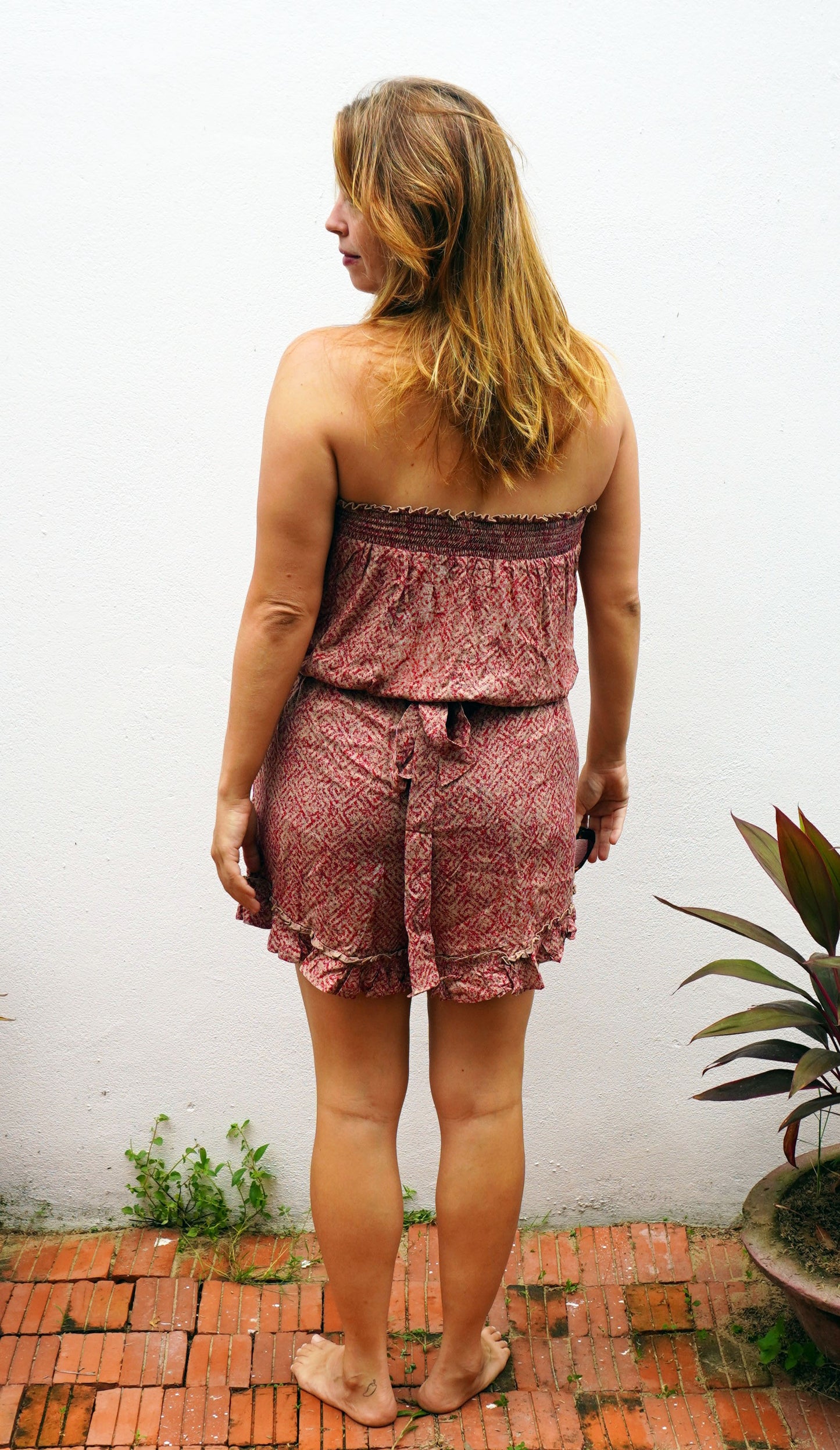 Woman wearing a pink patterned romper standing against a white wall with a plant in the background.