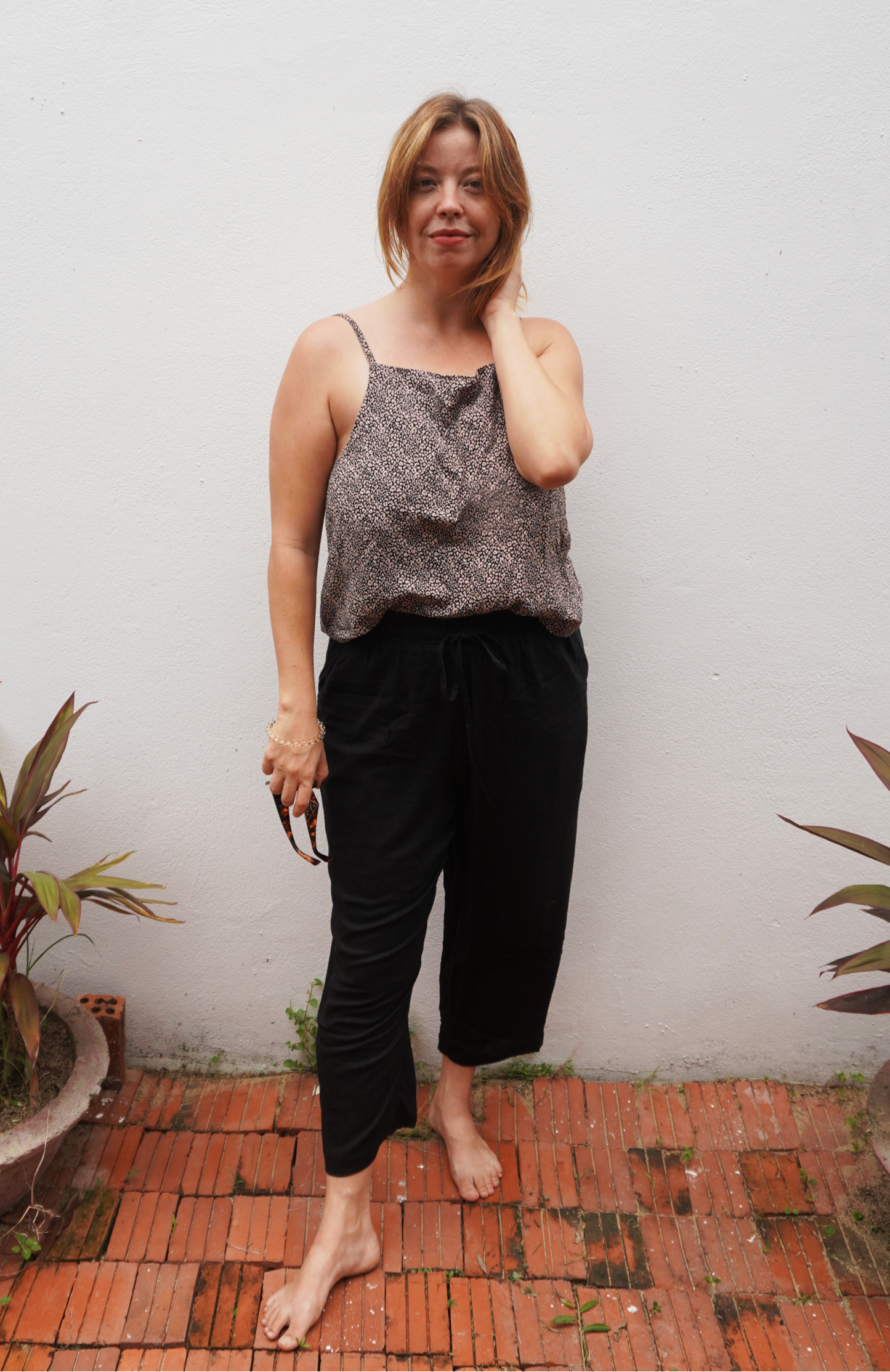 Woman wearing a leopard print patterned top and black pants standing on a brick floor with plants around.
