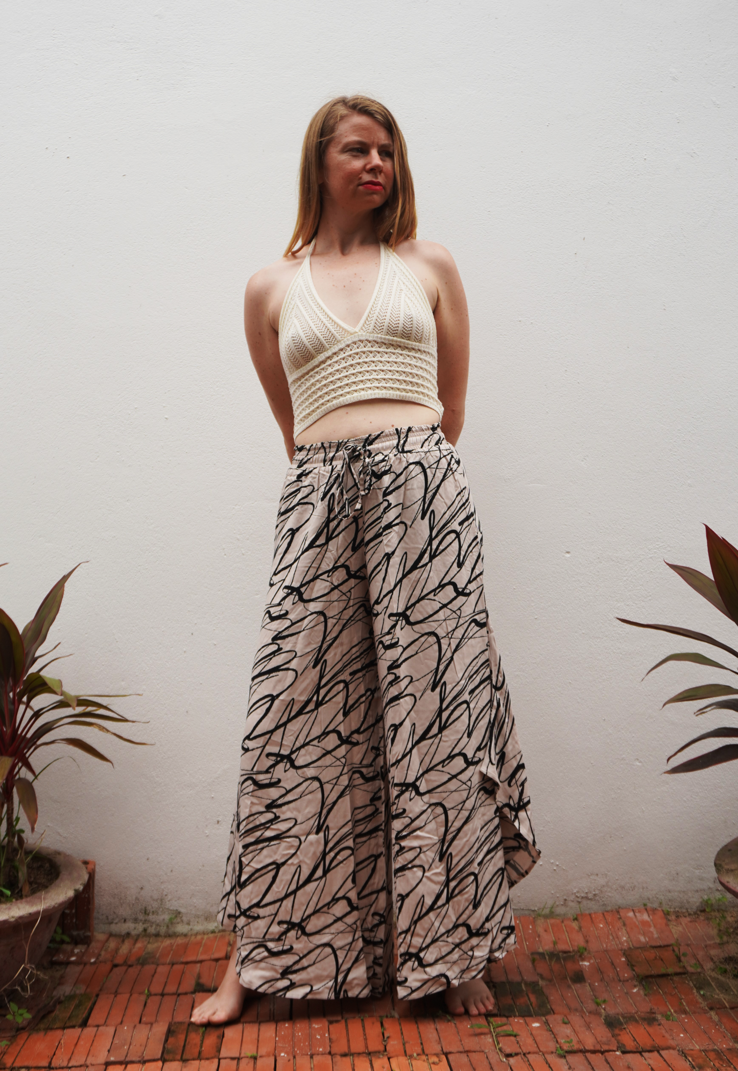 Woman wearing a white top and patterned pants standing against a plain wall with plants around.