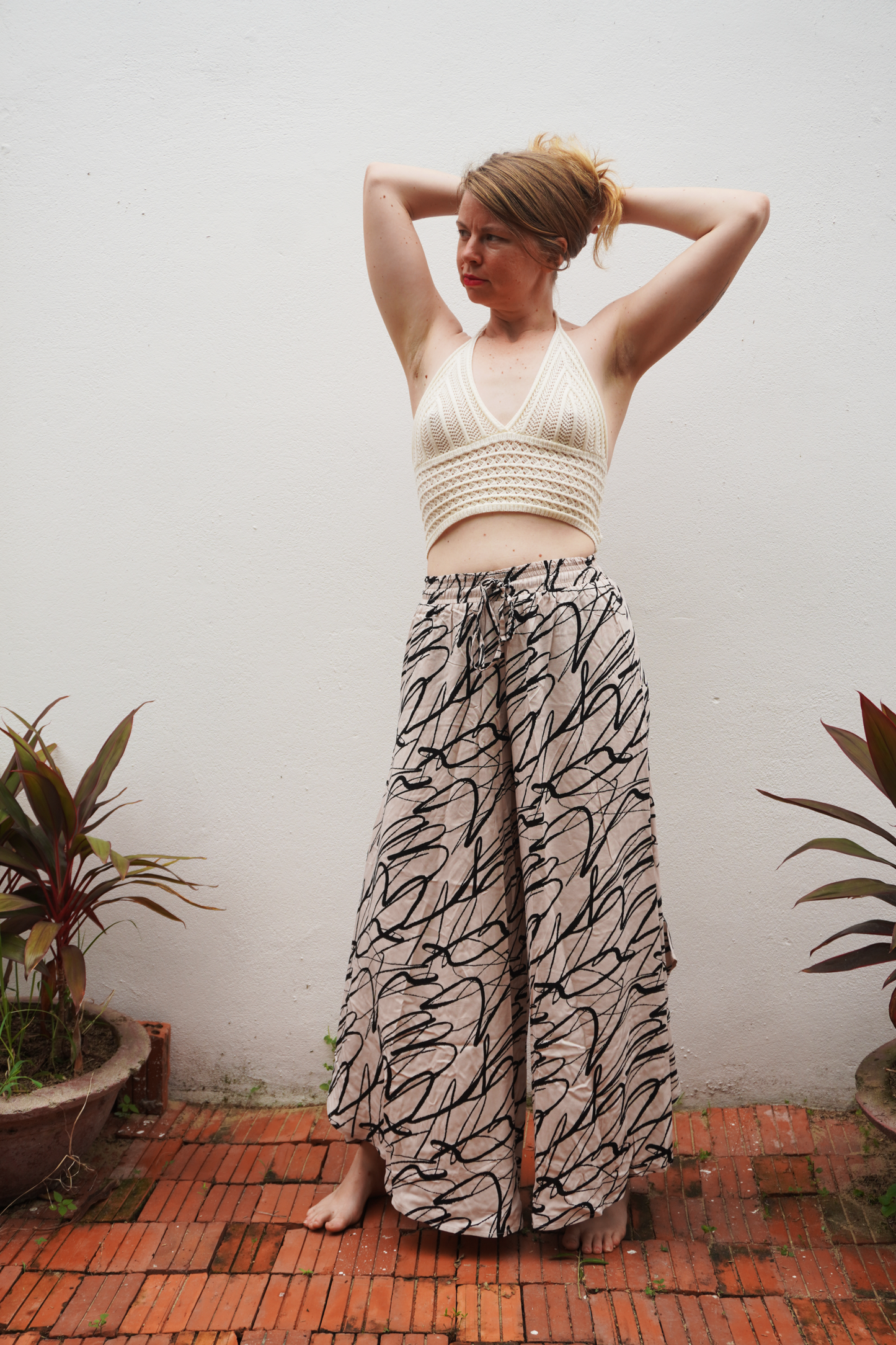 Woman wearing a white top and patterned pants standing against a white wall with plants around.