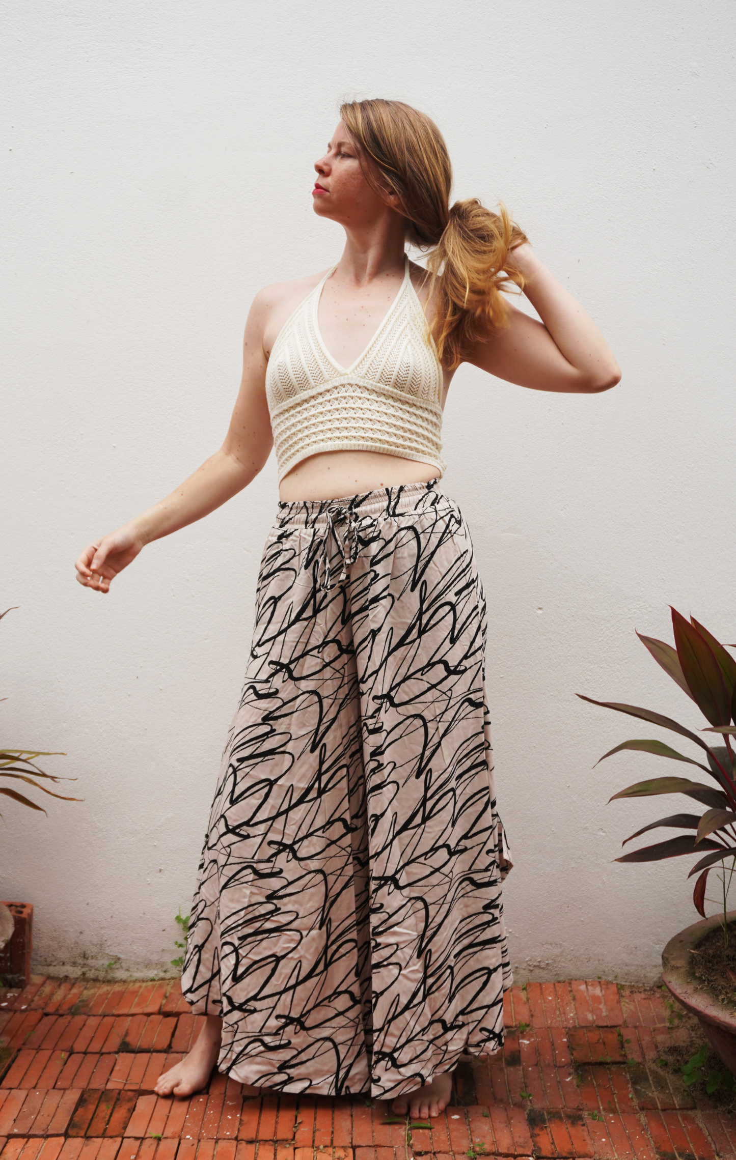 Woman wearing a white top and patterned skirt standing against a plain wall with plants around.