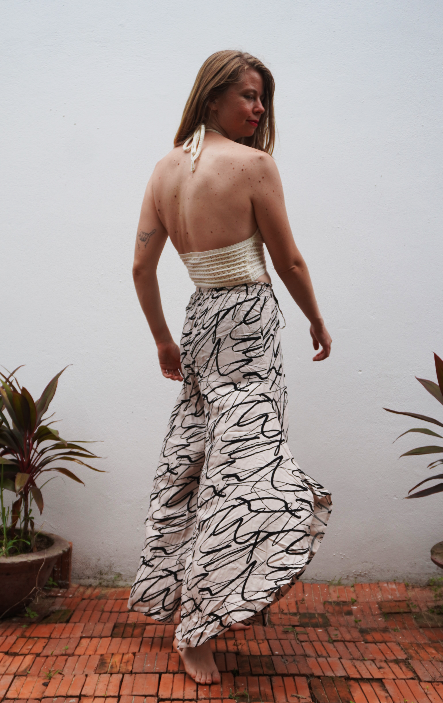 Woman wearing a white top and black and white patterned pants standing against a plain wall with plants around.