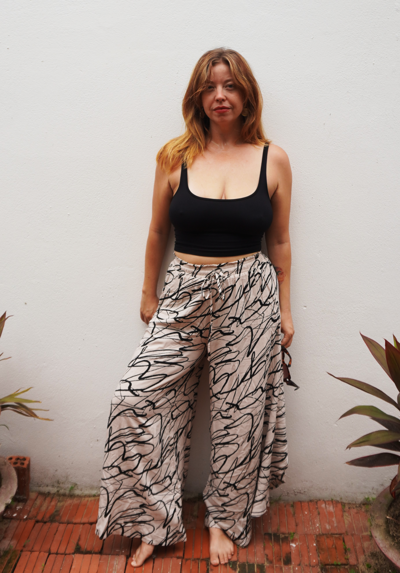 Woman wearing a black top and patterned pants standing against a white wall with plants around.