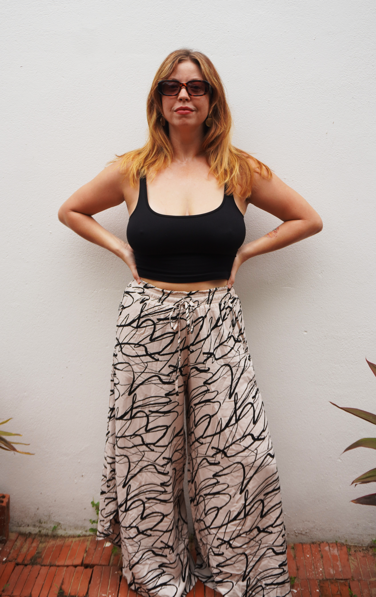 Woman wearing a black top and patterned pants against a white wall.