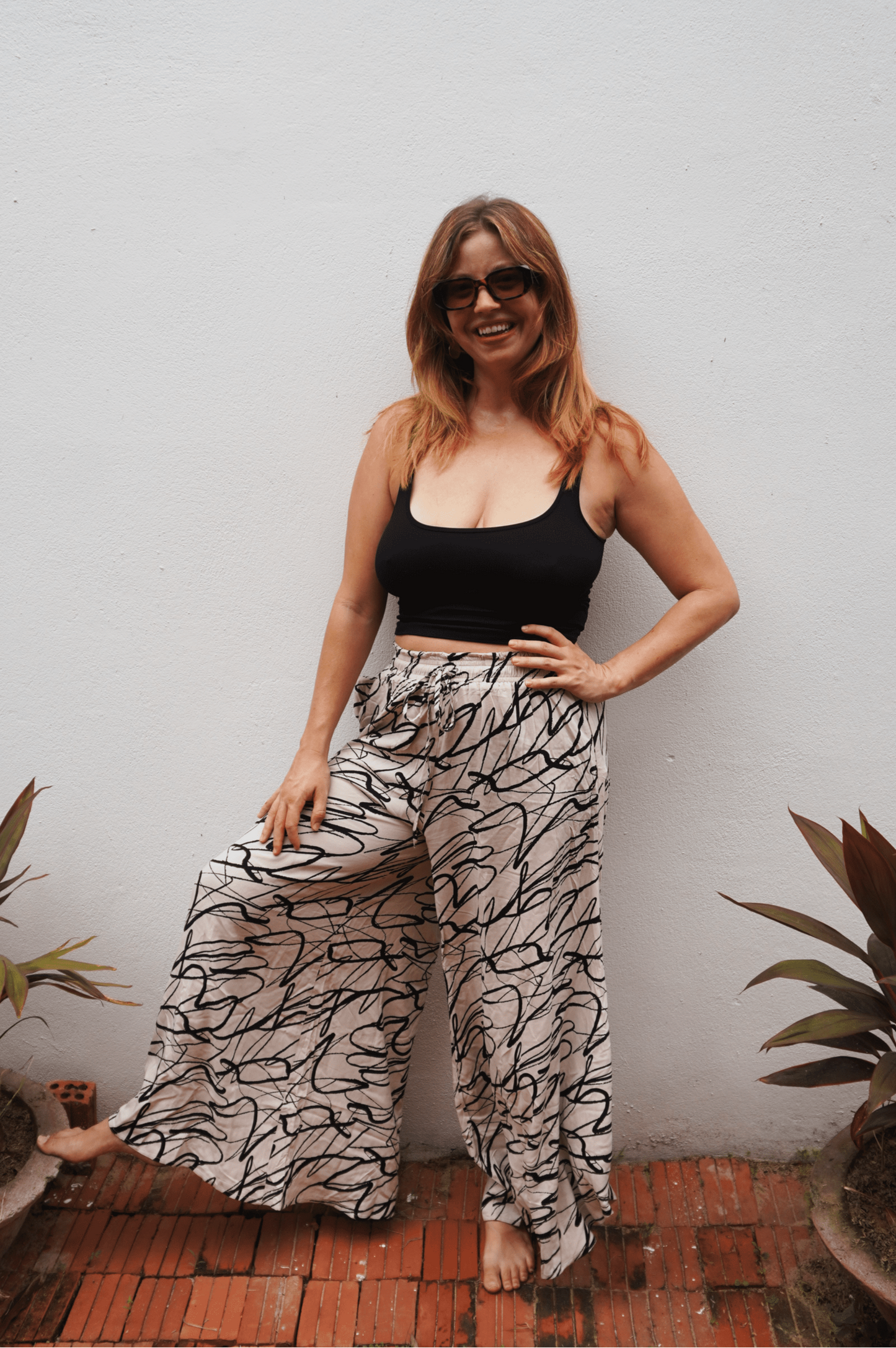 Woman wearing a black top and patterned pants standing against a white wall with plants around.