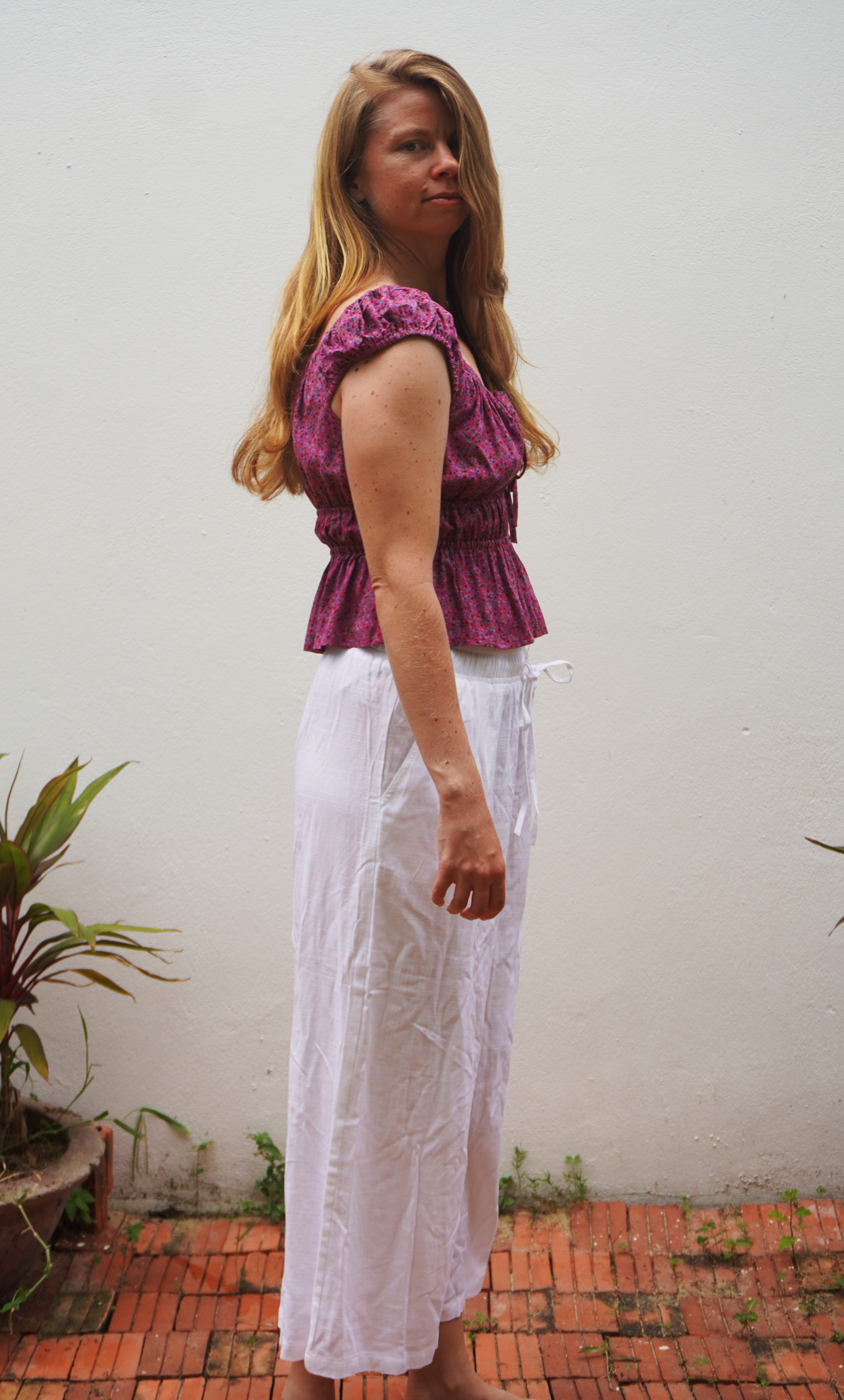 Woman wearing a purple top and white pants standing against a plain wall with plants.