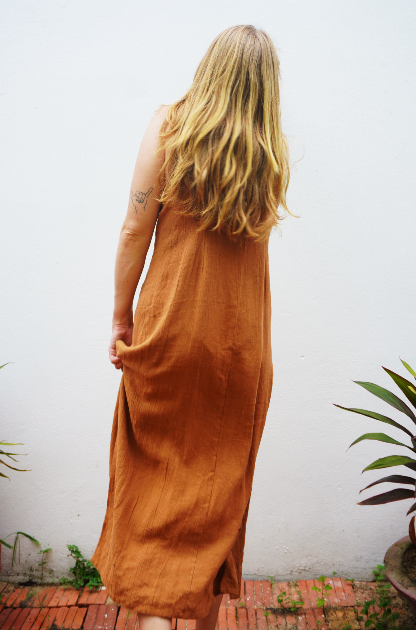 Person wearing a rust-colored dress standing against a white wall with plants around.