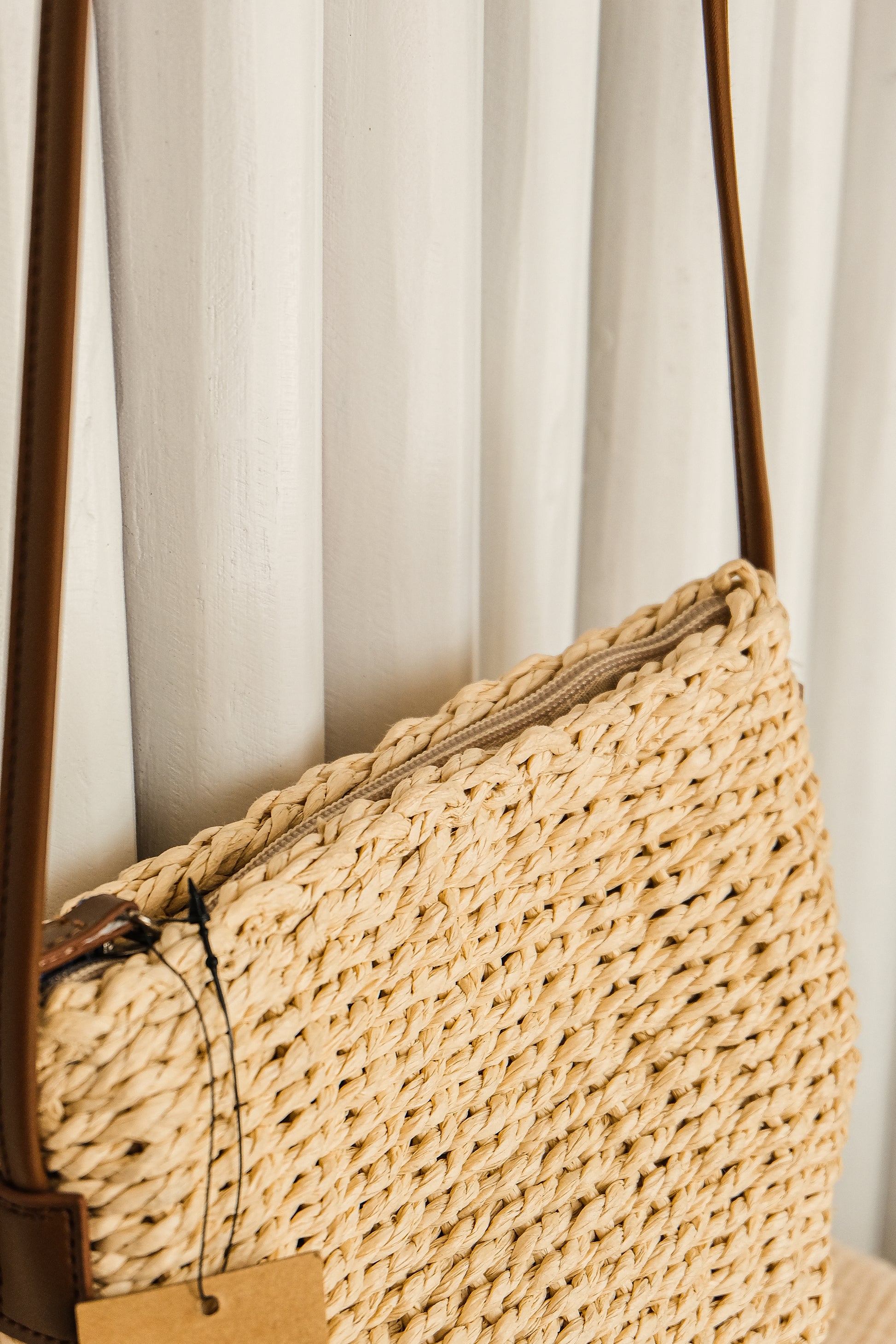 Woven straw handbag with brown leather strap against a white curtain background