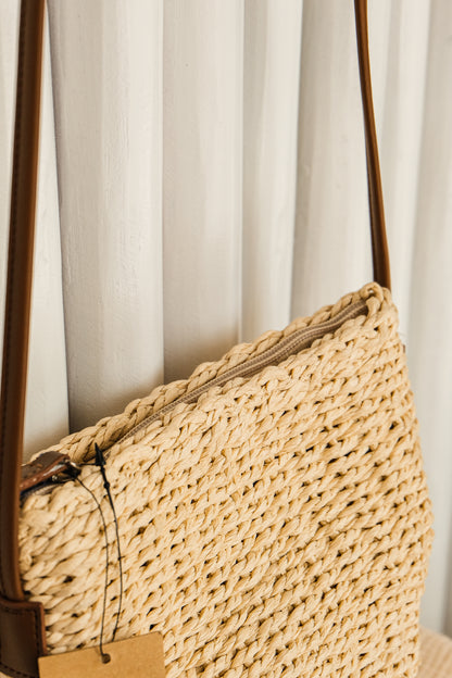 Woven straw handbag with brown leather strap against a white curtain background