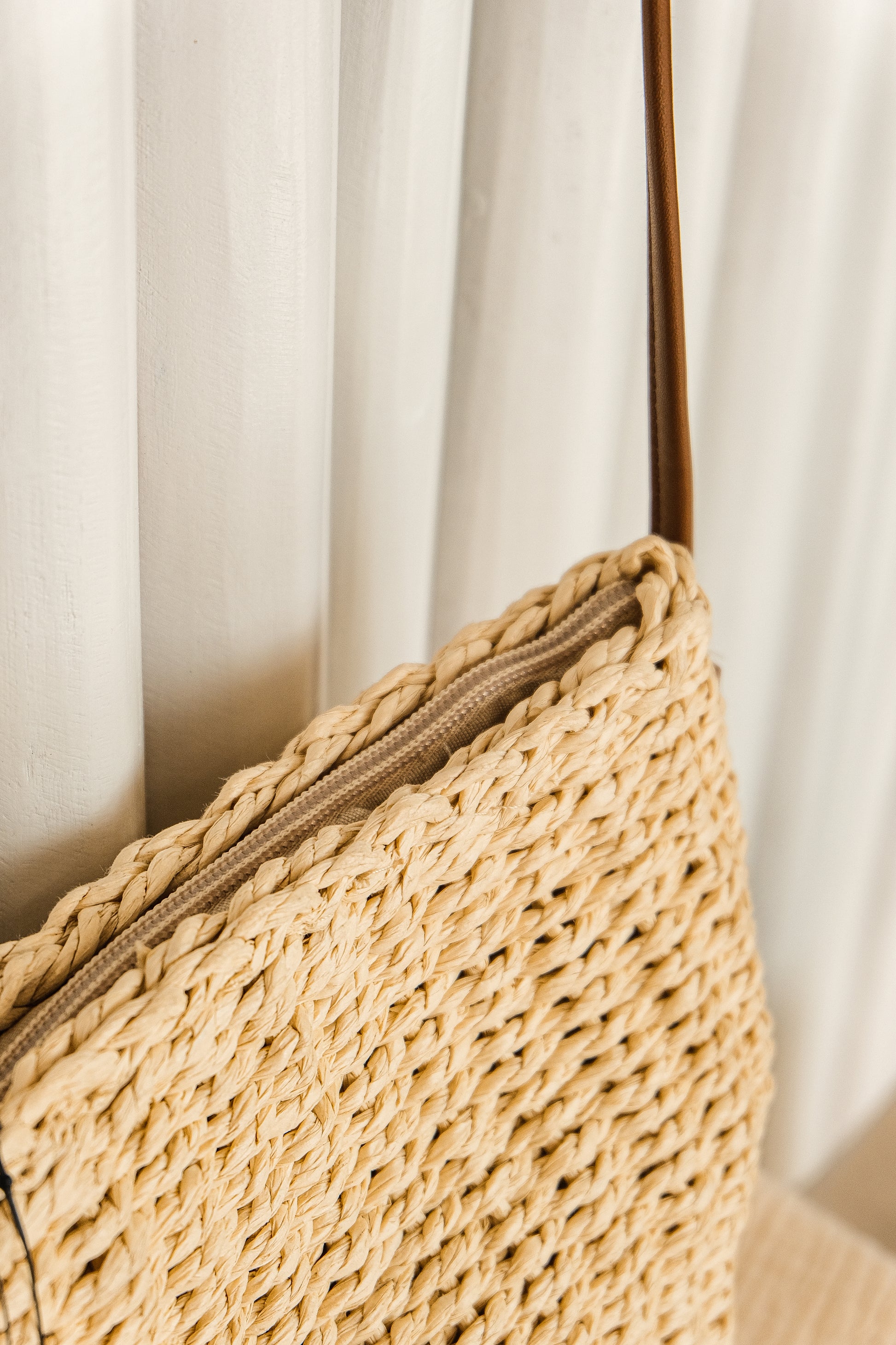 Close-up of a woven straw bag with a brown strap against a white curtain background