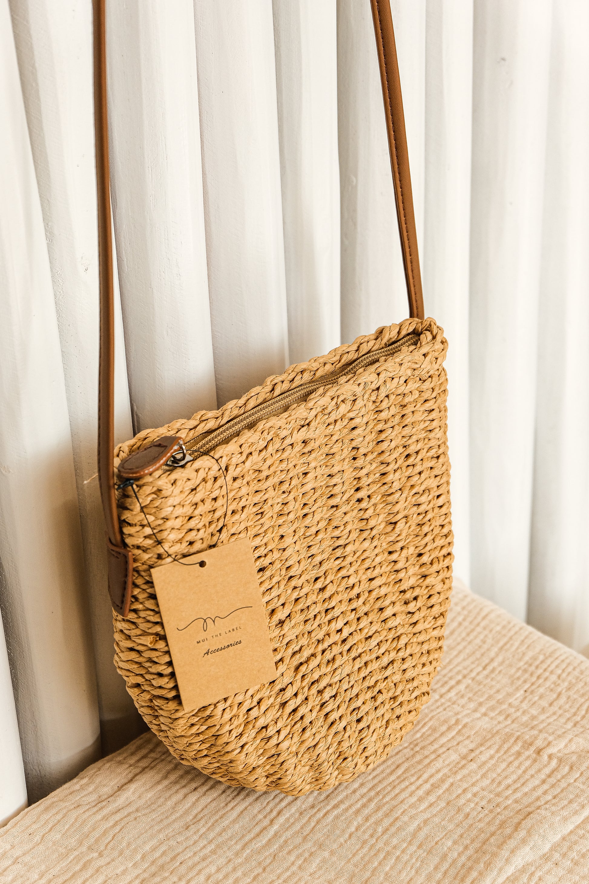 Woven beige handbag with a strap on a light wooden surface against a white curtain background