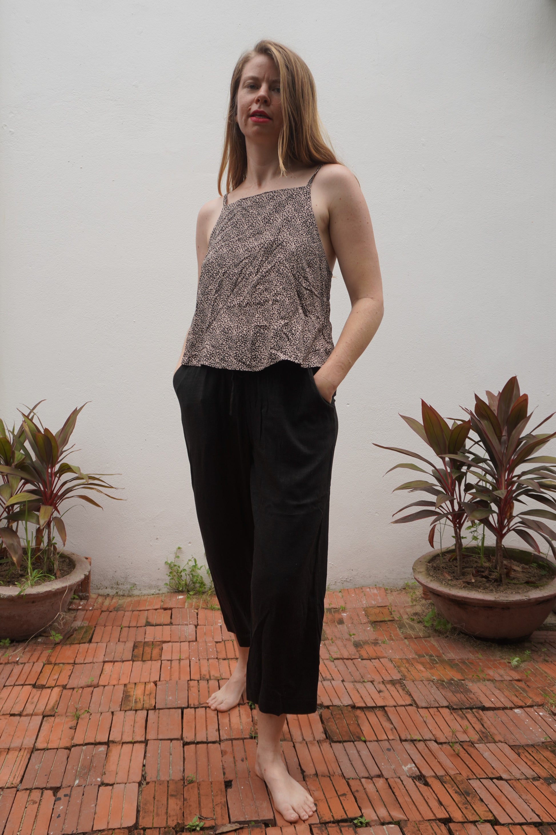 Woman wearing a patterned top and black pants standing on a brick patio with plants around.