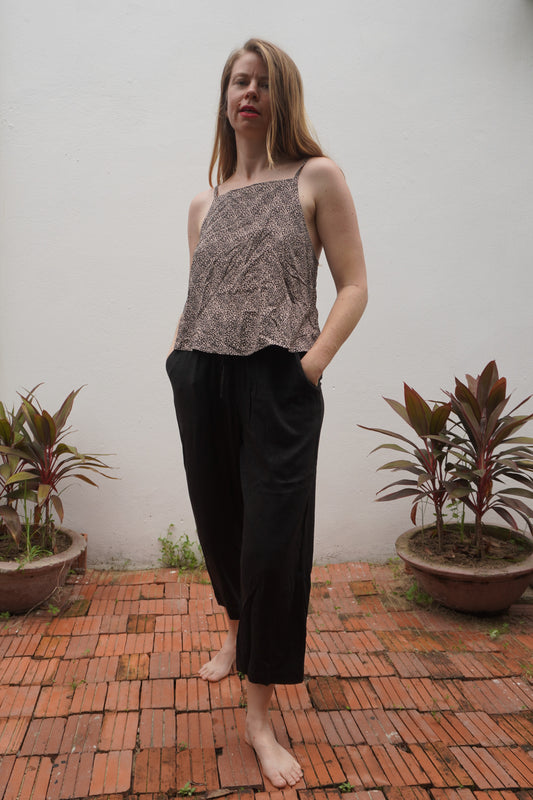 Woman wearing a patterned top and black pants standing on a brick patio with plants around.