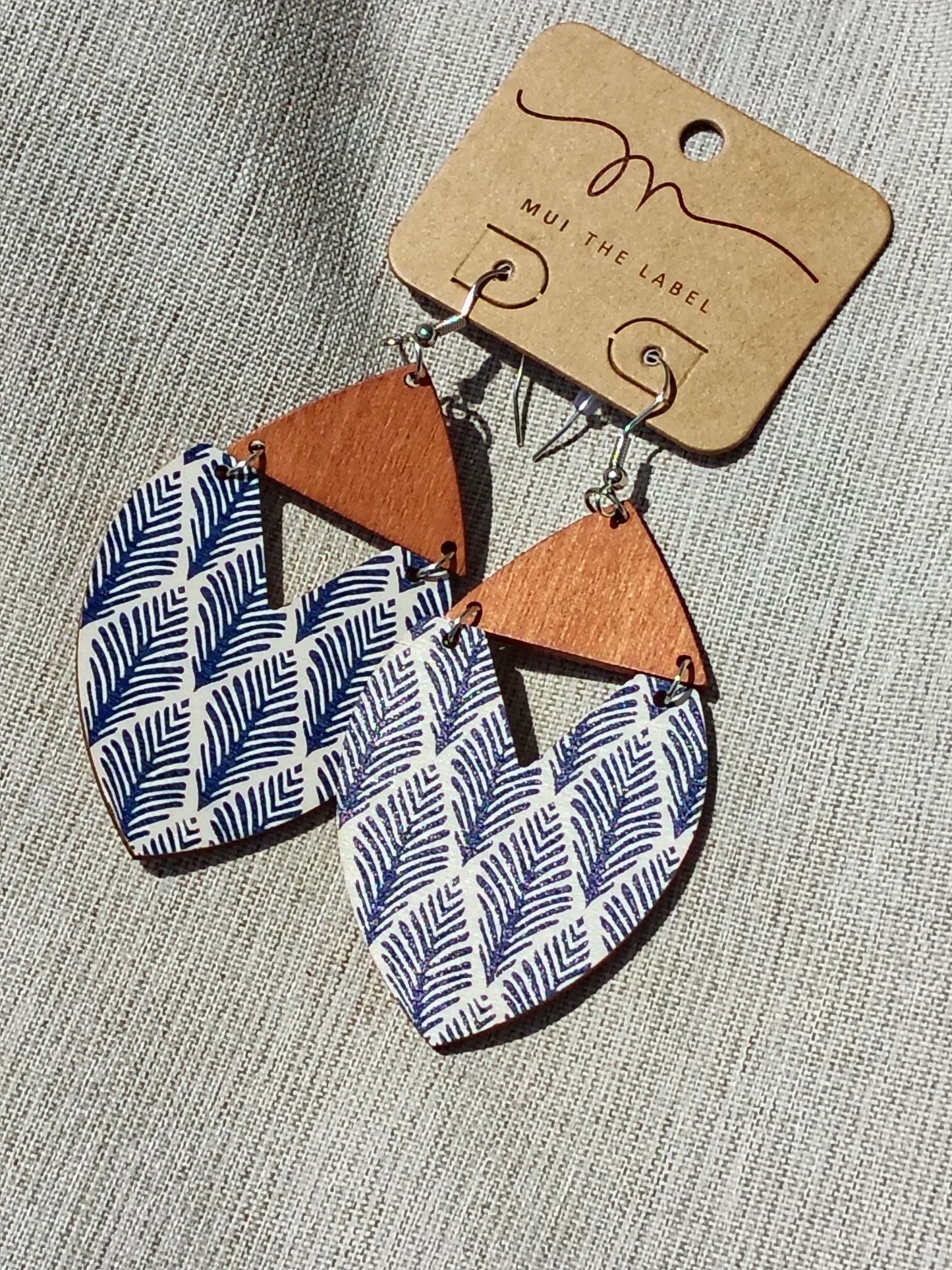 Earrings with blue and white patterned leaves and brown triangles on a gray textured background with 'Mui The Label' card.