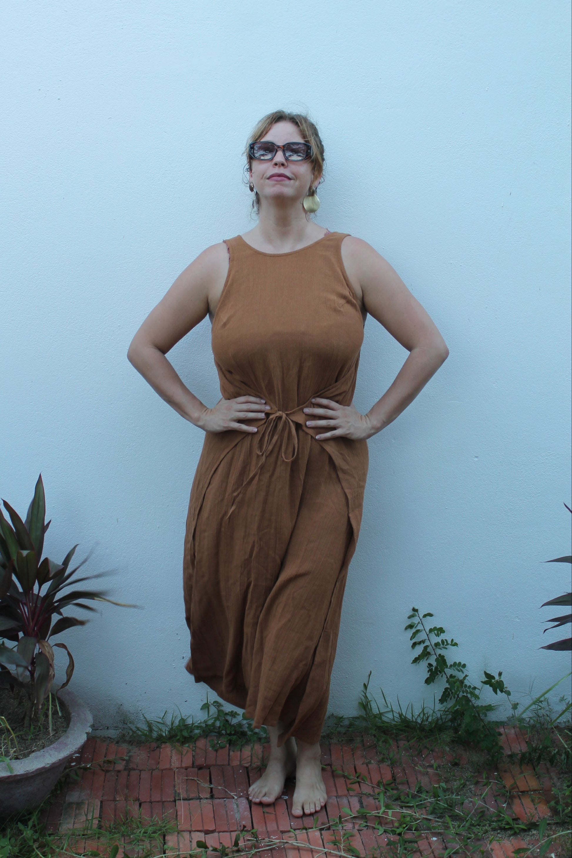Woman in a brown dress standing against a light blue wall with plants around.