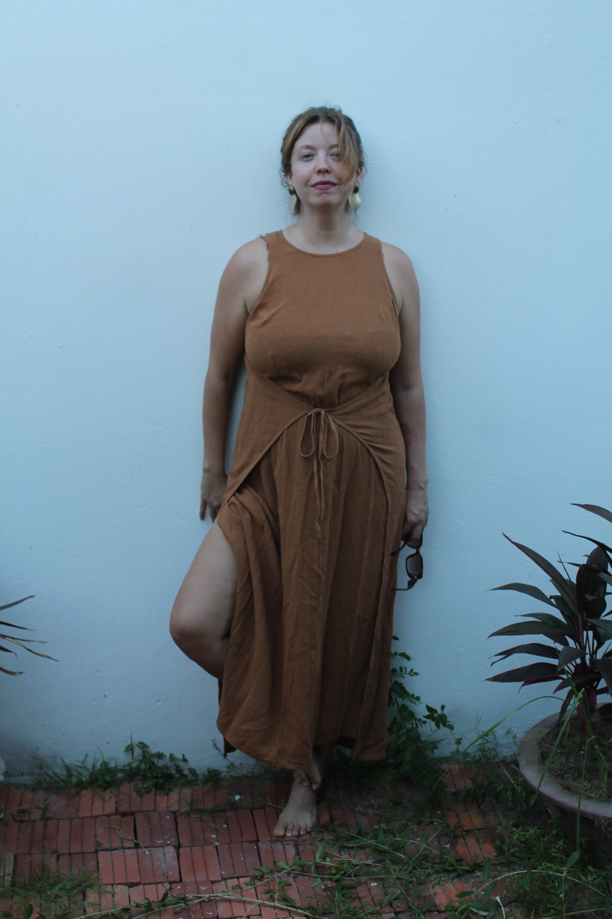 Woman wearing a brown dress standing against a light blue wall with plants around.