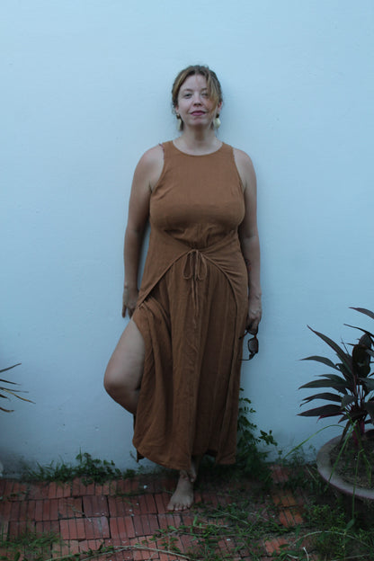 Woman wearing a brown dress standing against a light blue wall with plants around.