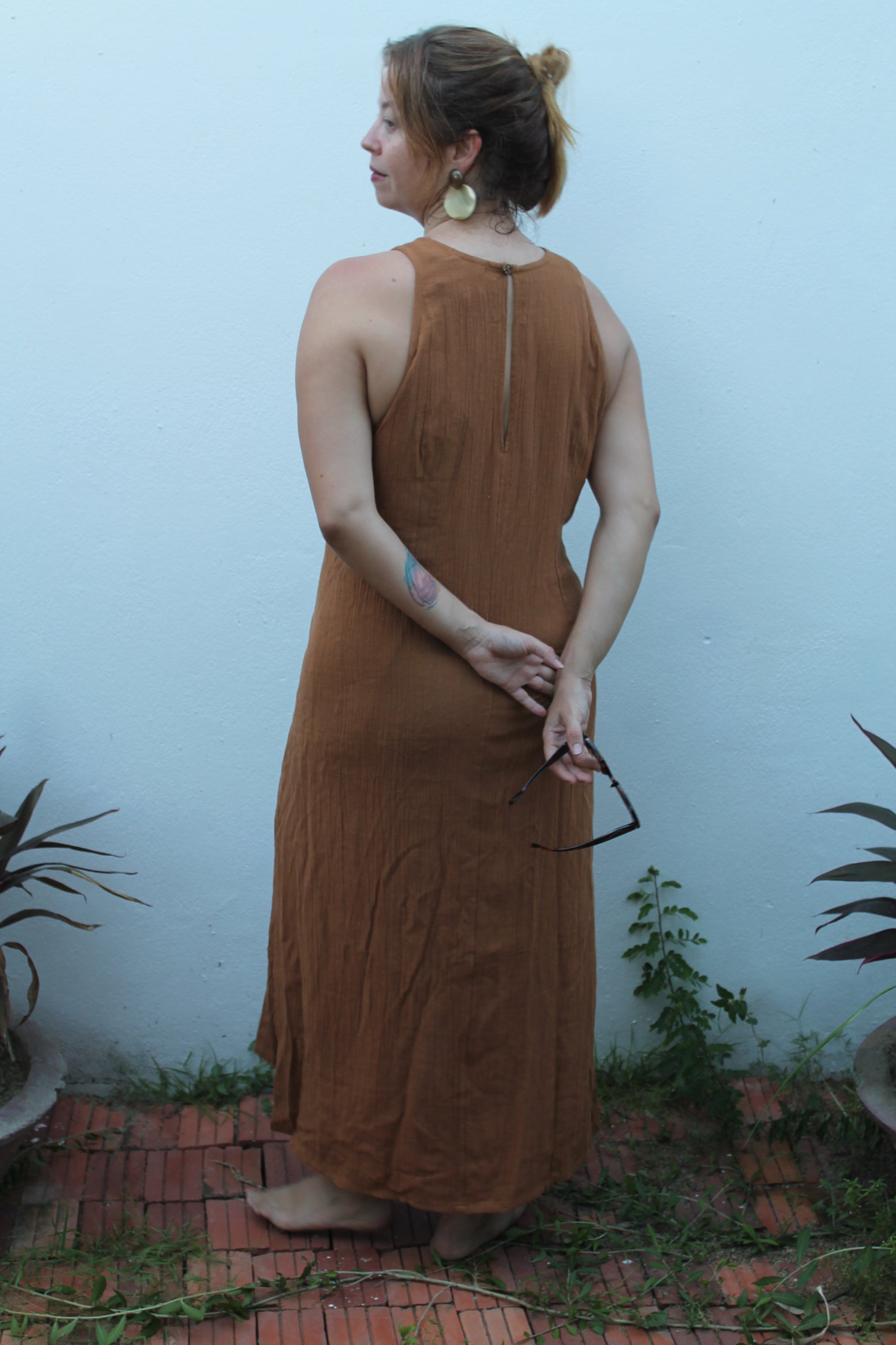 Woman in a brown dress standing against a light blue wall with plants around.