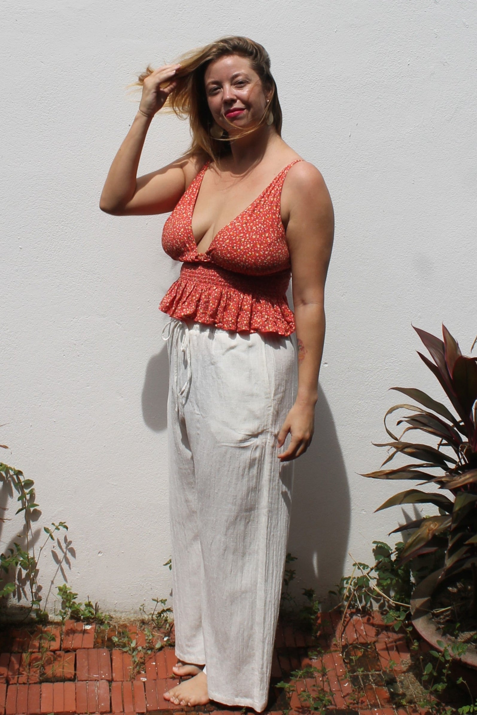 Woman wearing a red top and white pants standing against a white wall with plants around.