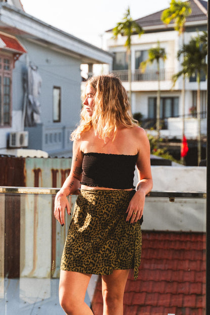 Woman in a black top and leopard print skirt standing on a rooftop with buildings and plants in the background.