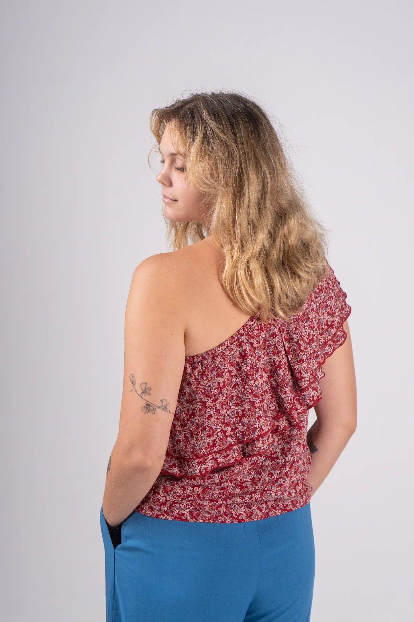 Woman wearing a red floral one-shoulder top and blue pants on a white background