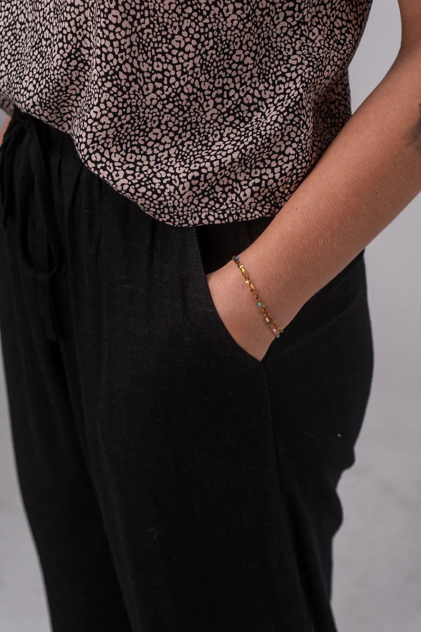 Person wearing a patterned top and black pants with a bracelet on a neutral background