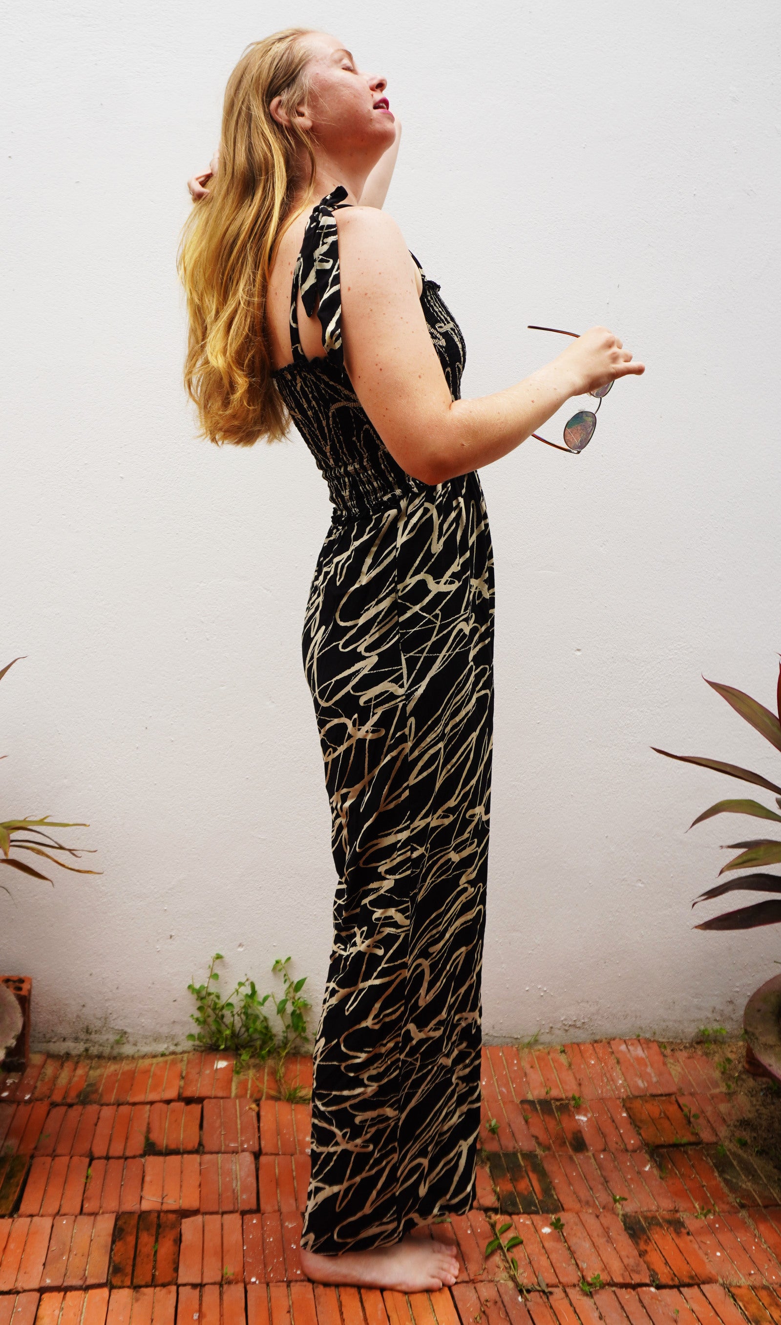 Woman wearing a black and cream patterned jumpsuit standing on a brick patio with plants in the background.