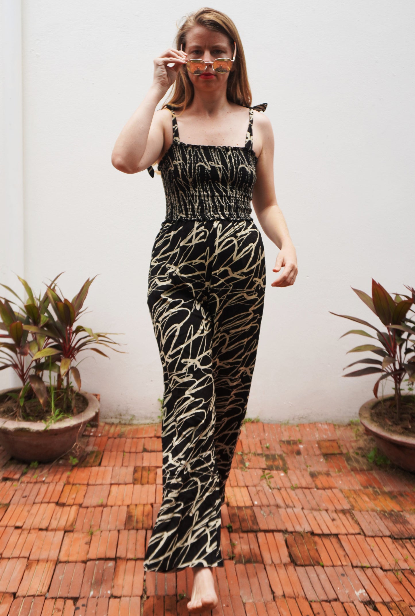Woman wearing a black and white patterned outfit standing on a brick patio with plants in the background.