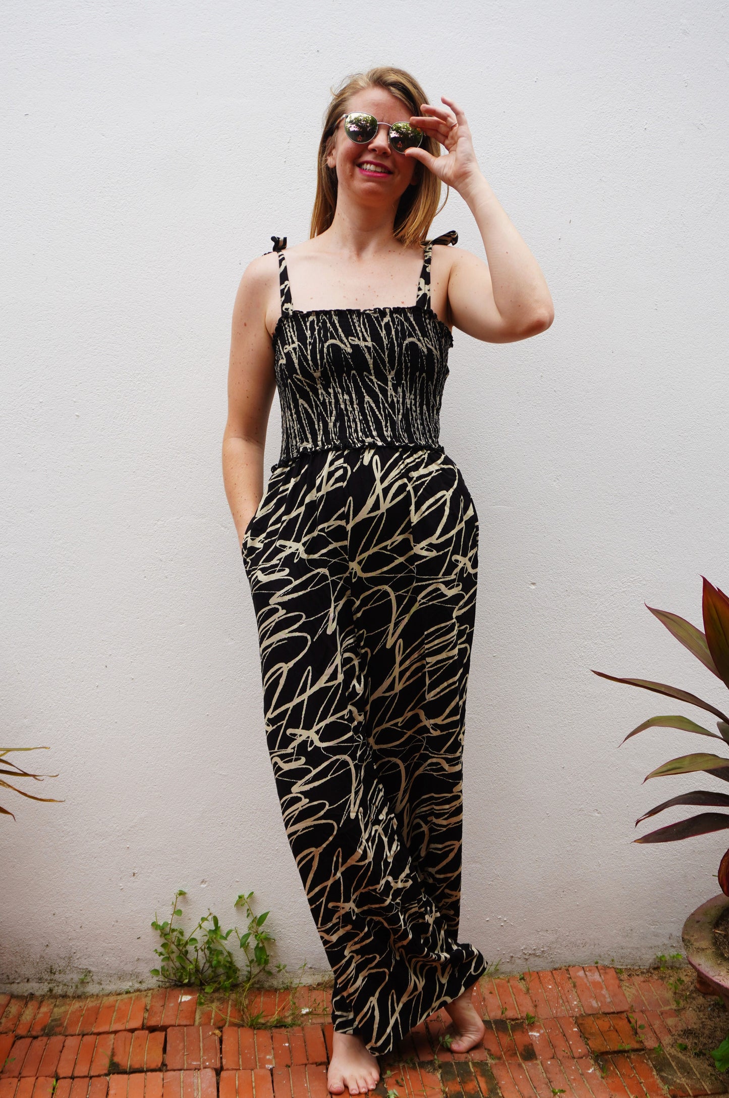 Woman wearing a black and cream patterned dress standing against a white wall with plants around.