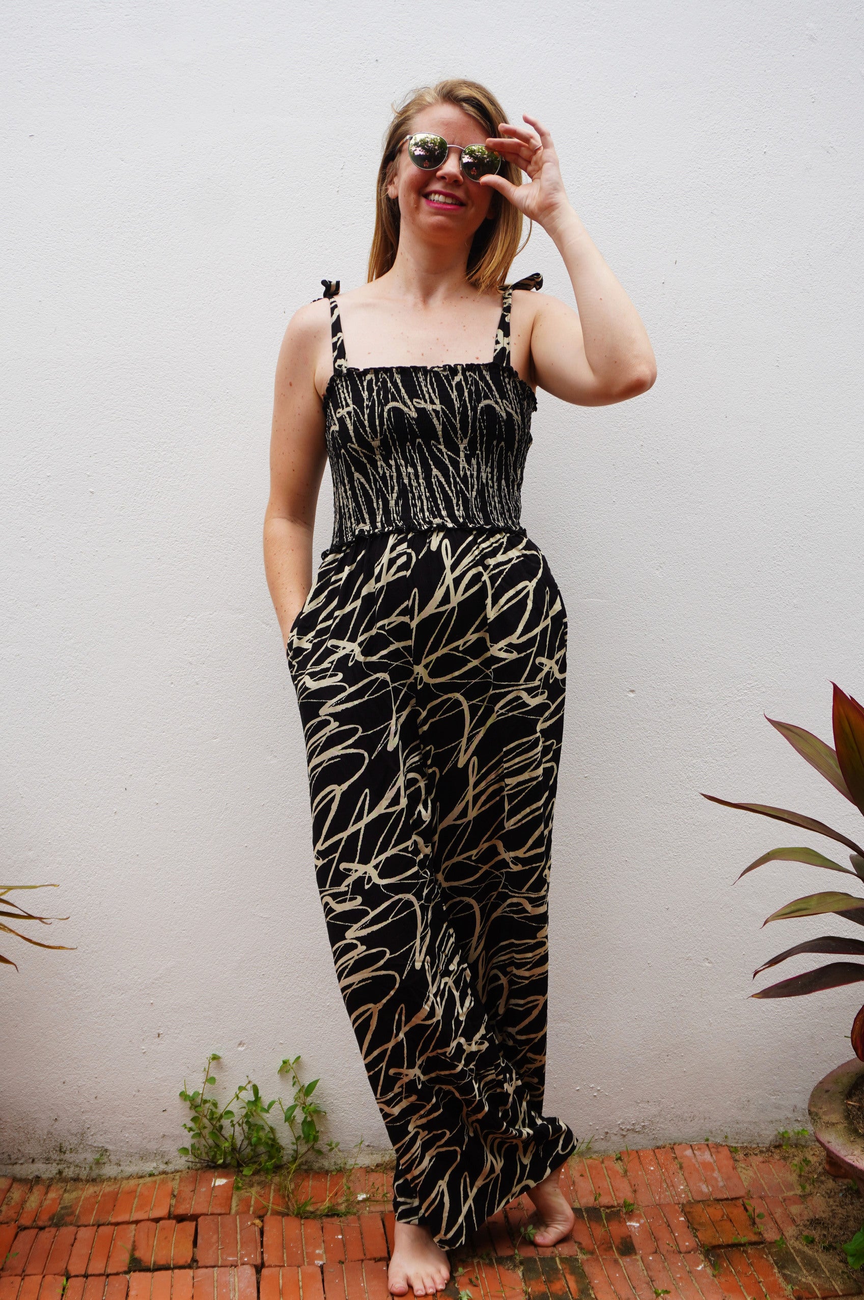 Woman wearing a black and cream patterned dress standing against a white wall with plants around.