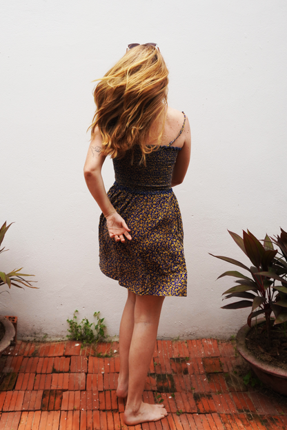 Woman in a purple and yellow floral patterned dress standing on a red brick floor with plants around.