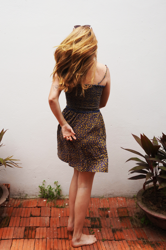 Woman in a purple and yellow floral patterned dress standing on a red brick floor with plants around.