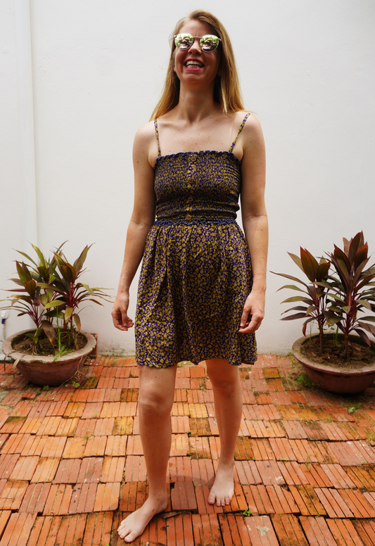 Woman wearing a purple and yellow floral patterned dress standing on a wooden floor with plants in the background