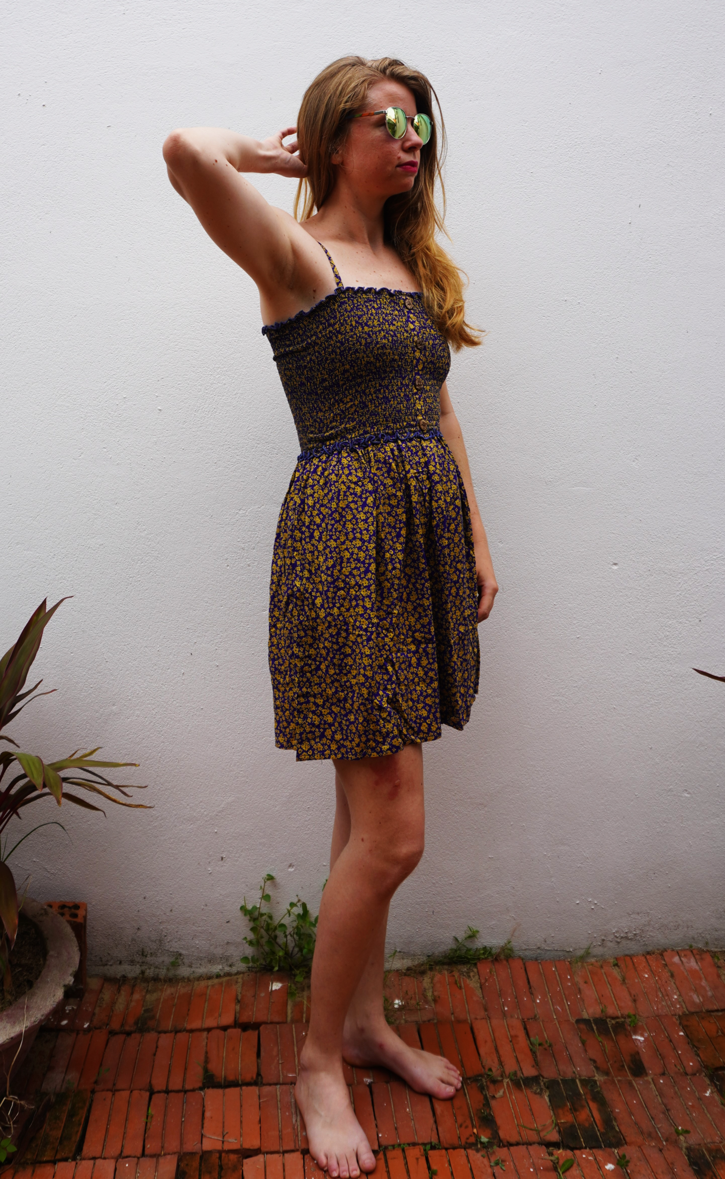 Woman in a purple and yellow floral  patterned dress standing on a brick floor with a white wall background