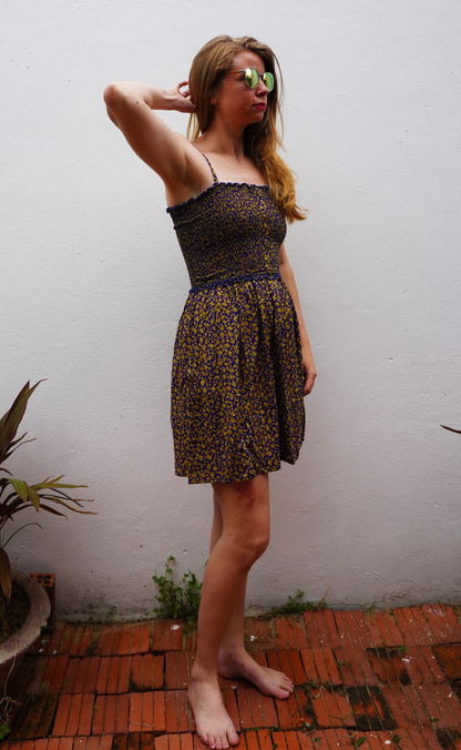 Woman in a purple and yellow floral  patterned dress standing on a brick floor with a white wall background