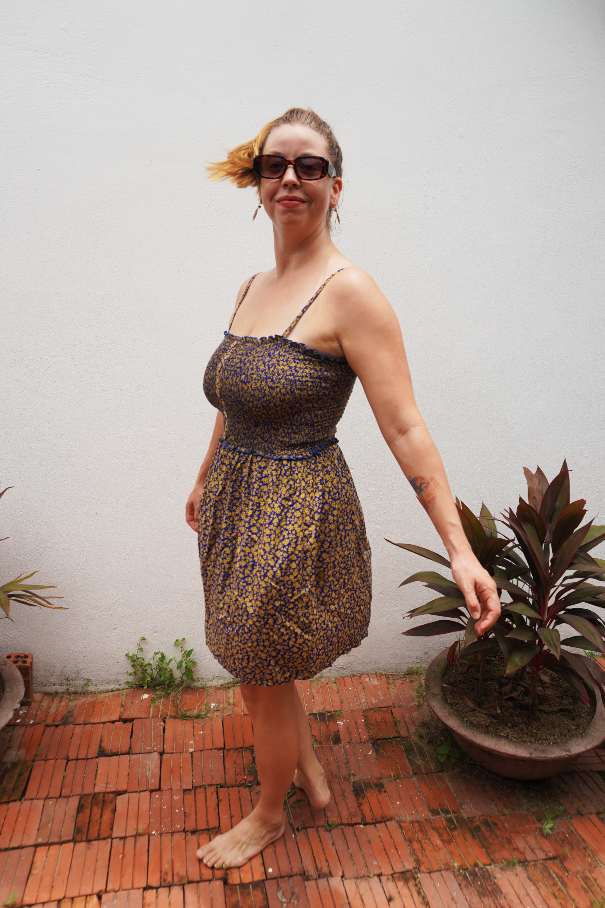 Woman in a patterned dress standing outdoors with a white wall and potted plant in the background