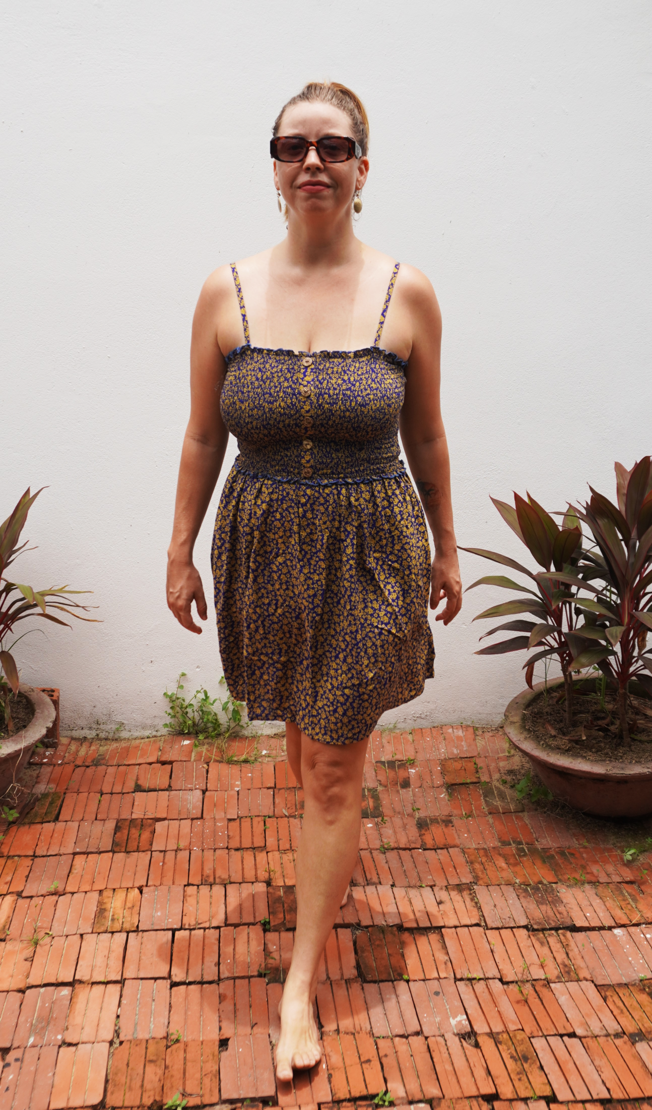 Woman wearing a purple and yellow floral patterned dress standing on a brick patio with plants in the background