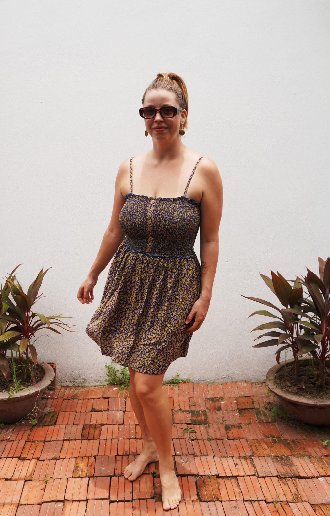Woman in a purple and yellow floral patterned dress standing on a brick patio with plants around.