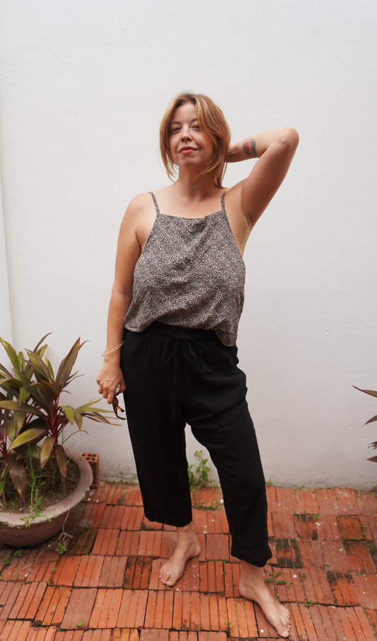 Woman wearing a patterned top and black pants standing on a brick floor with plants around.