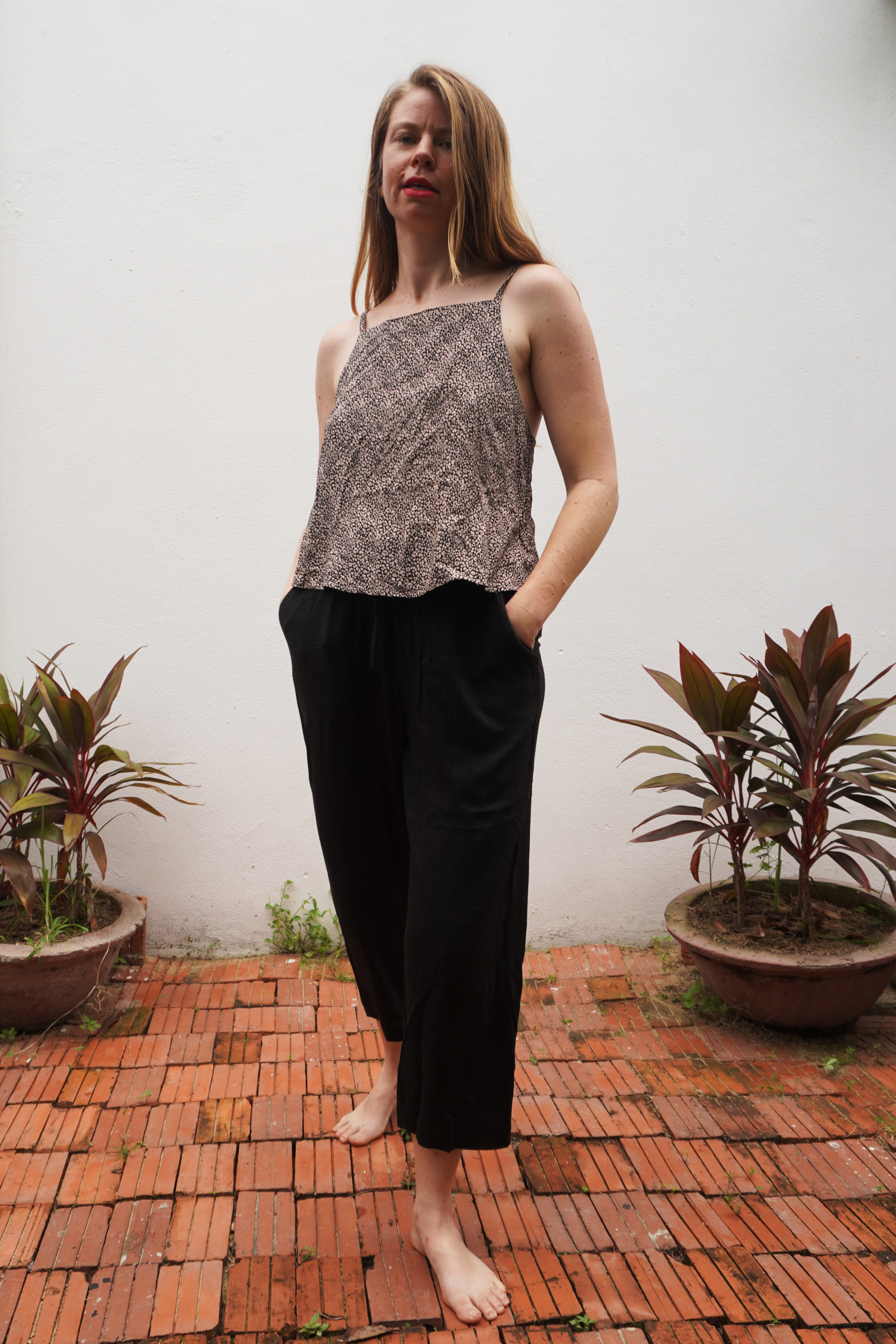 Woman wearing a leopard print patterned top and black pants standing on a brick patio with plants around.