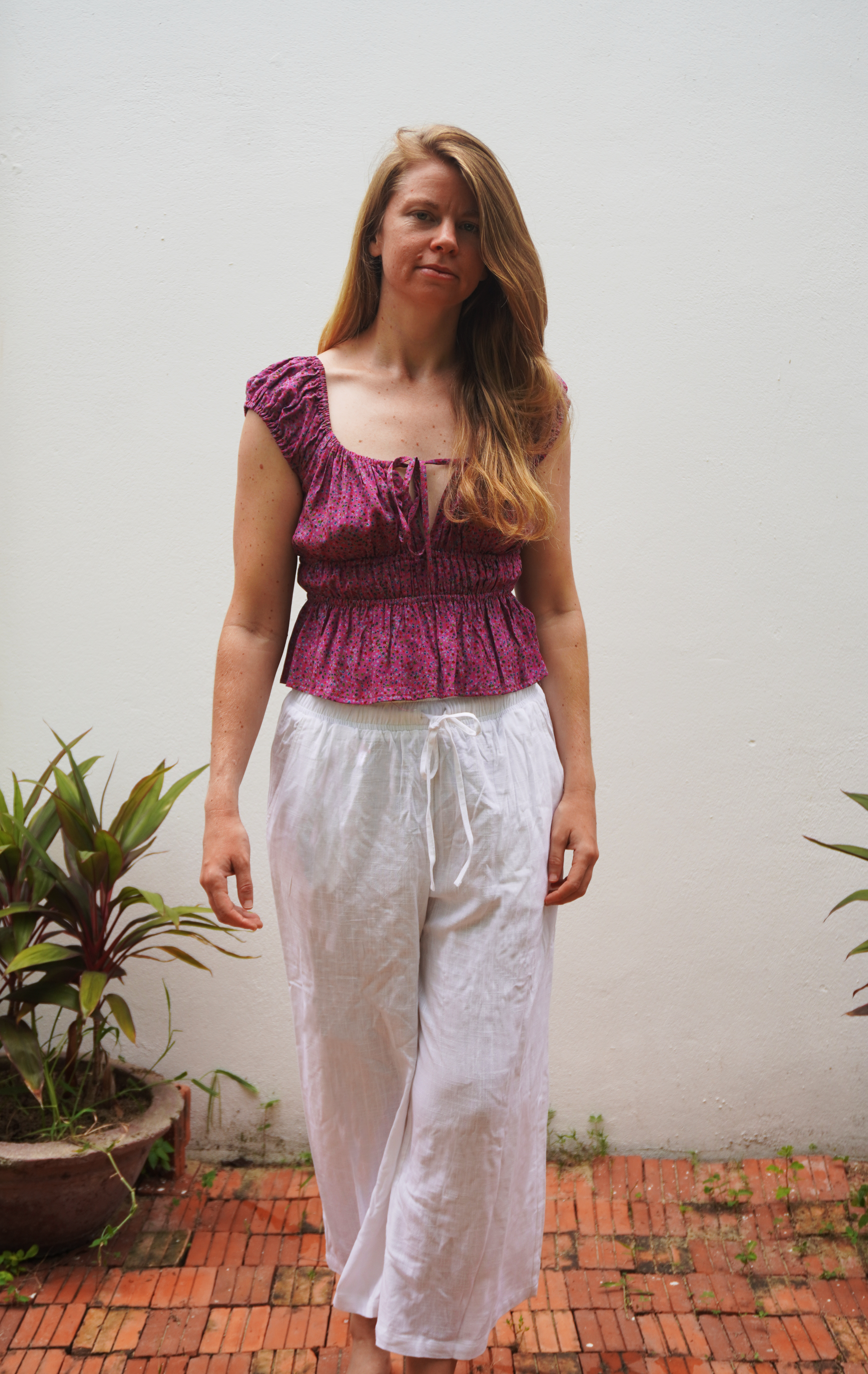 Woman wearing a purple top and white pants standing outdoors with plants in the background
