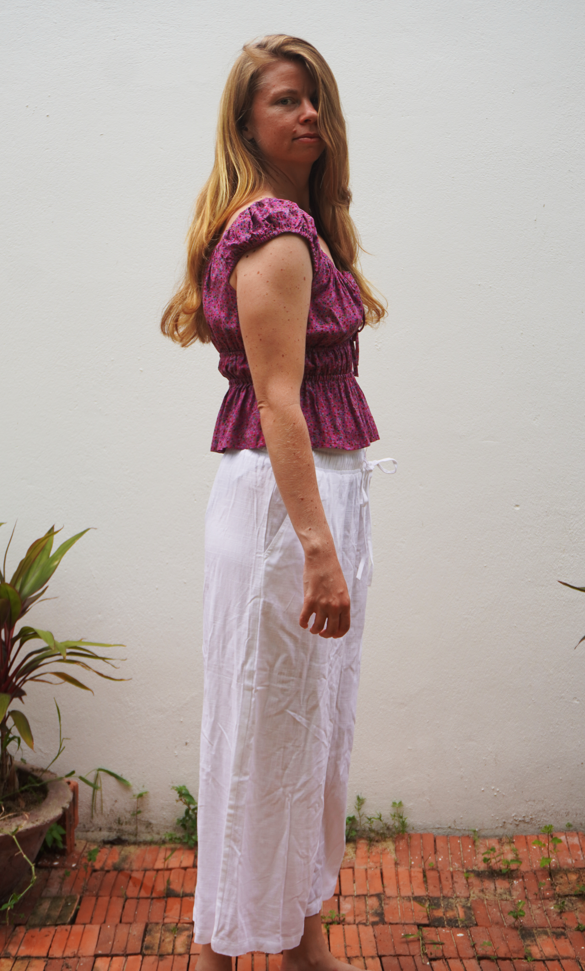 Woman wearing a purple top and white pants standing against a plain wall with plants.