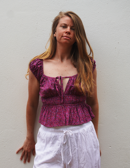 Woman wearing a purple patterned top and white pants against a plain background
