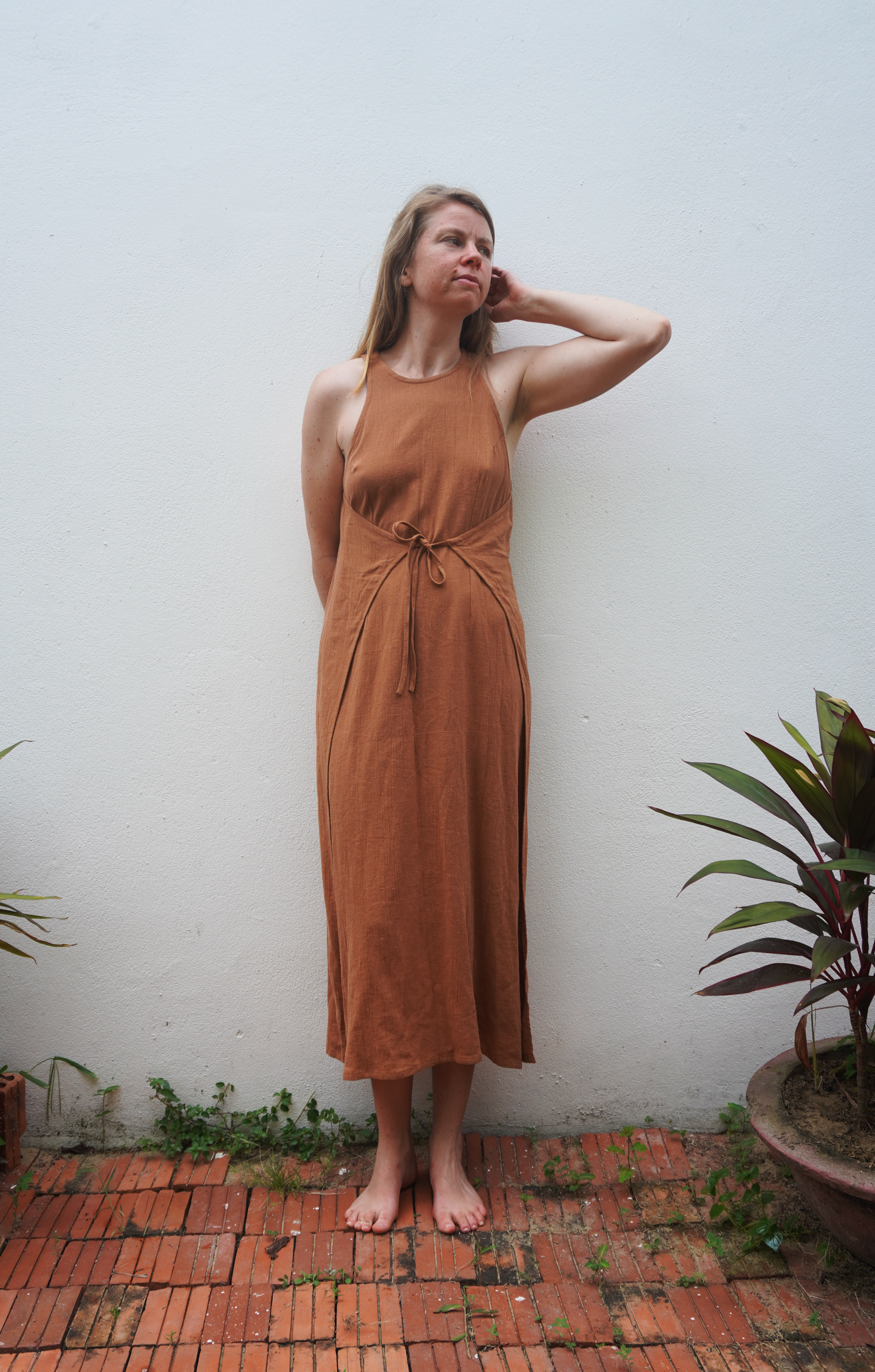 Woman in a brown dress standing against a white wall with plants around.