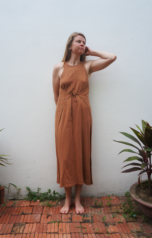 Woman in a brown dress standing against a white wall with plants around.