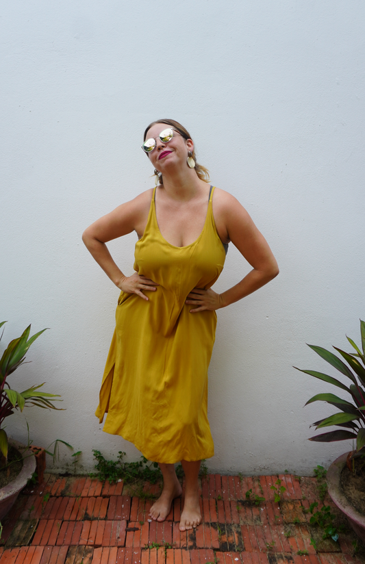 Woman in a yellow dress standing against a white wall with plants around.