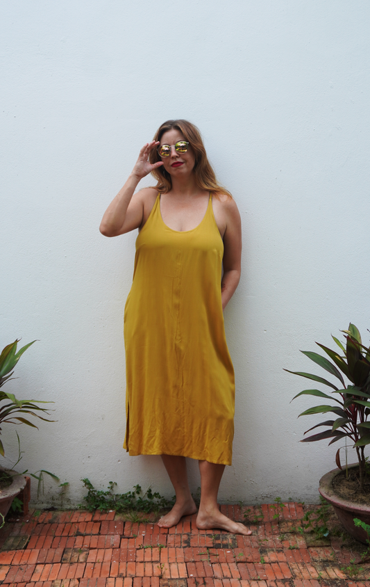 Woman wearing a yellow dress standing against a white wall with plants around.