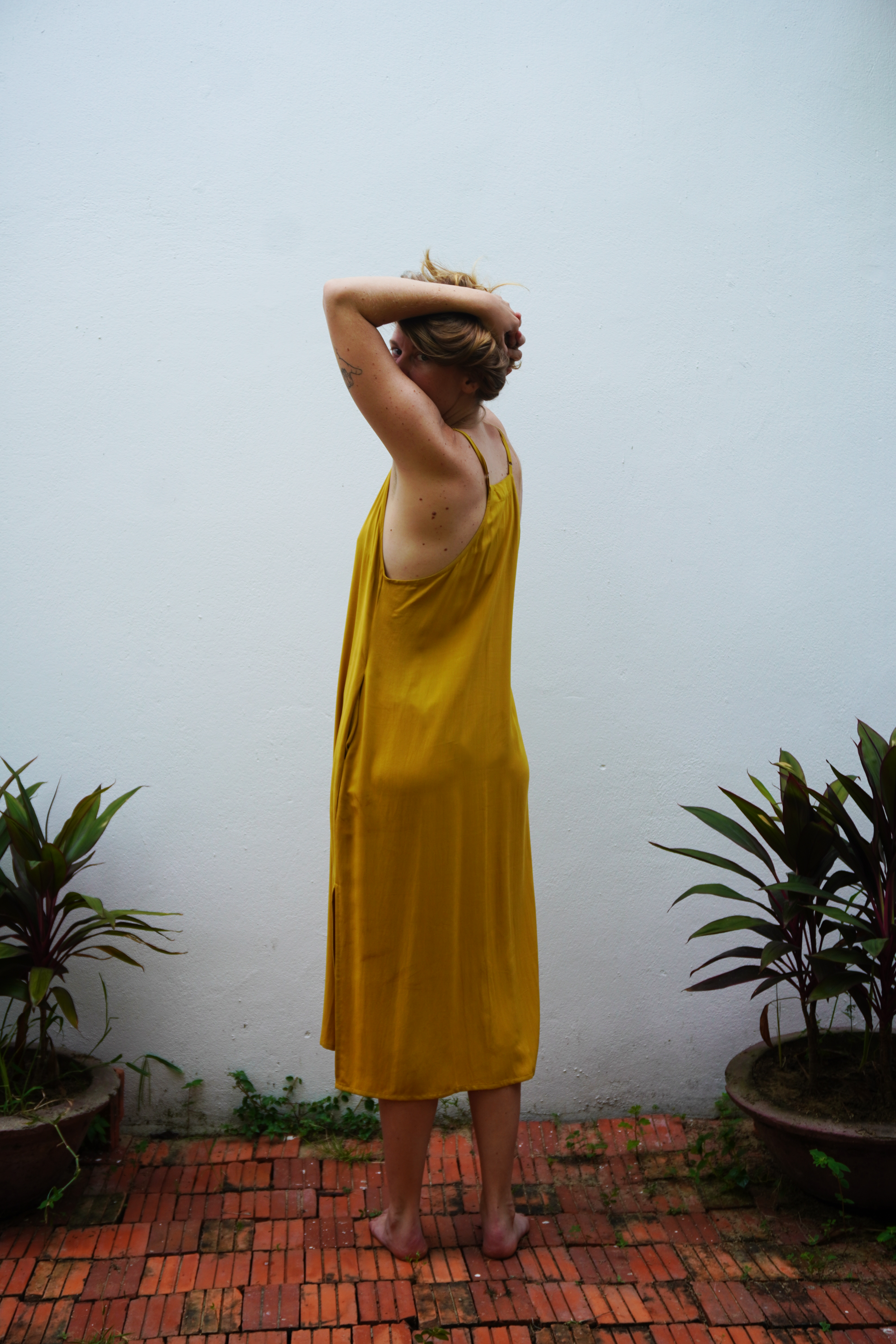 Person wearing a yellow dress standing in a garden with plants and a white wall.
