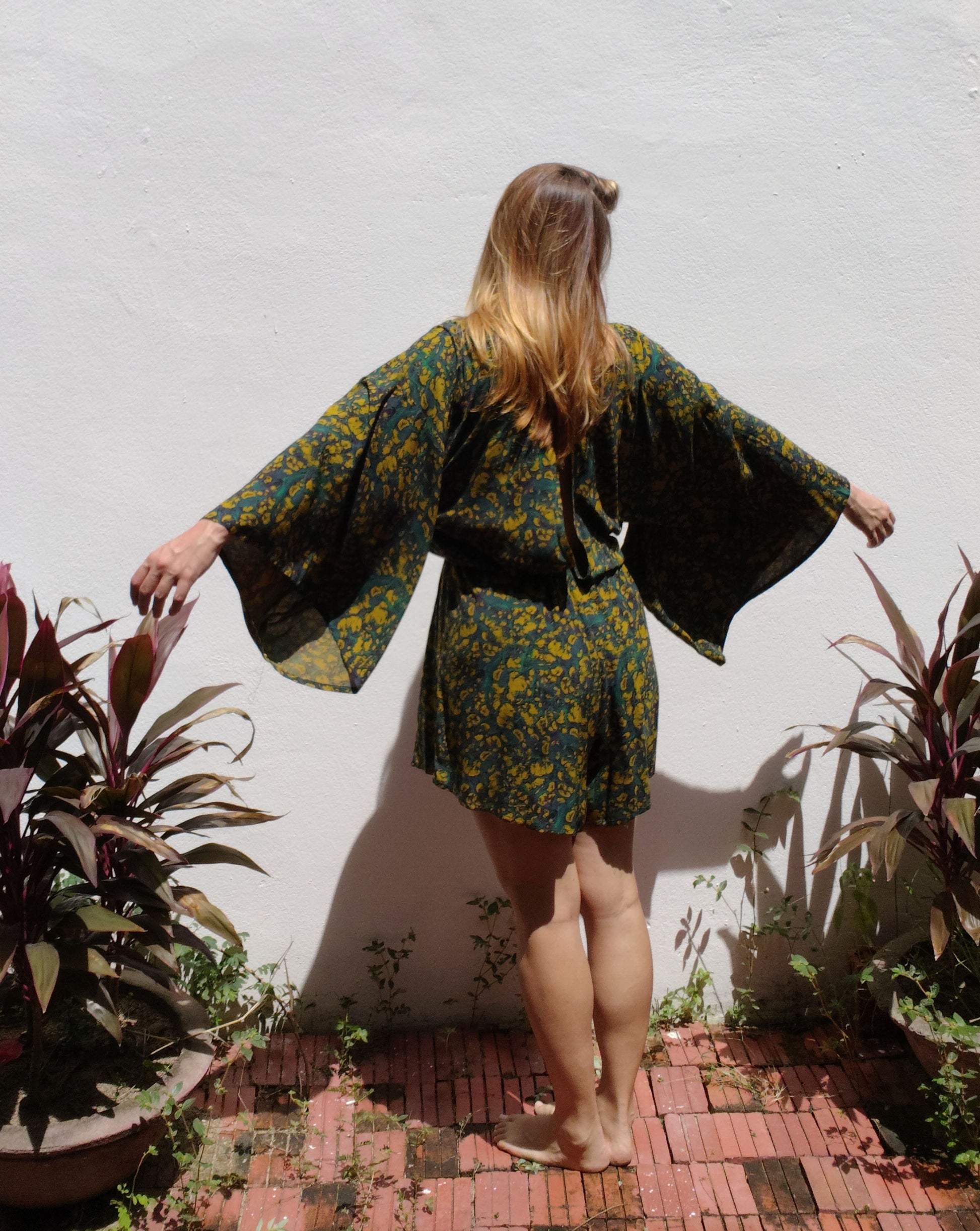 Woman in a green peacock printed romper standing in a garden with plants around.