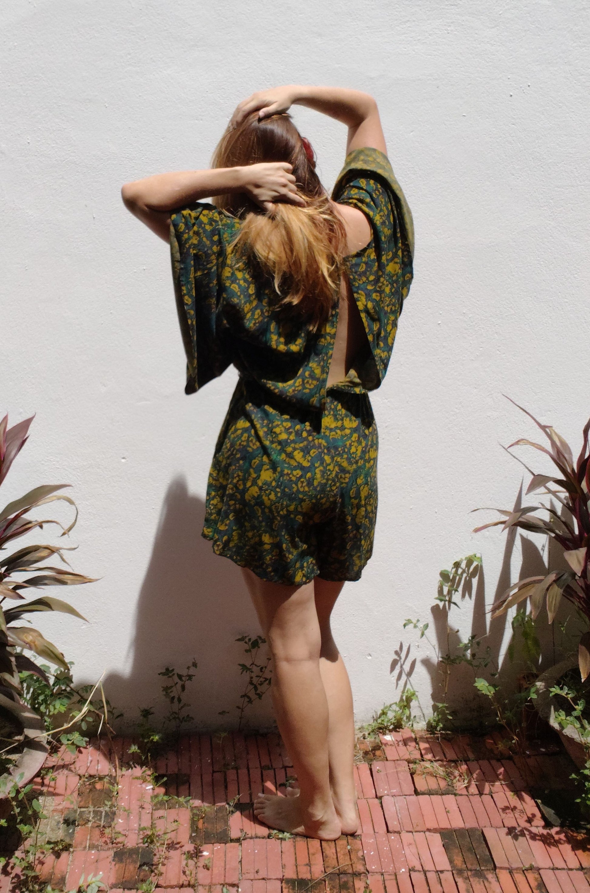 Person wearing a green floral jumpsuit standing against a white wall with plants around.